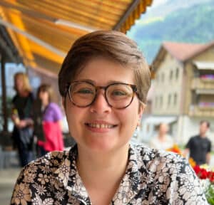 A white woman in her late 20s with short brown hair and round glasses wearing a black-and-white floral men's button-up.