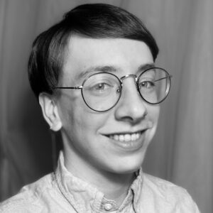 A photo of Oliver Dougherty, a white androgynous person wearing round glasses, a button-down, and a single dangling earring