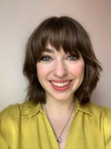 Headshot of a white woman with shoulder-length brown hair and a fringe smiling directly into the camera. She's wearing a light green silky shirt and has pink lipstick and rosy cheeks.