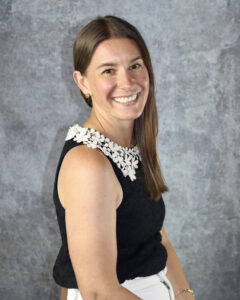 Brown hair woman sitting in front of grey backdrop. White collar shirt and white pants with black lace top.