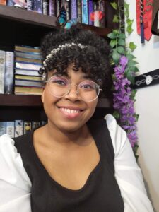 received_698065382045859 A Photo of Leah Moss. She is smiling and sitting in front of a flower-adorned bookshelf.
